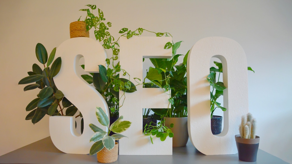White 3D letters spelling SEO surrounded by hanging and potted green plants on a gray surface - Photo by Lukas Müller on Unsplash